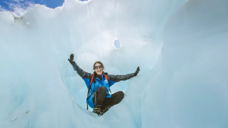 A smiling hiker sits inside a natural ice chamber on Fox Glacier.