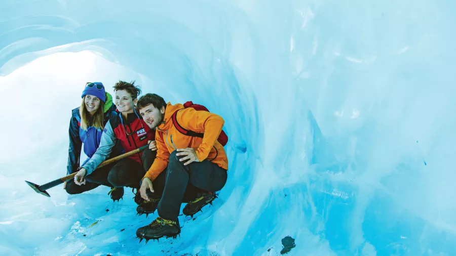 Three hikers crouch inside a glowing blue ice cave at Fox Glacier.