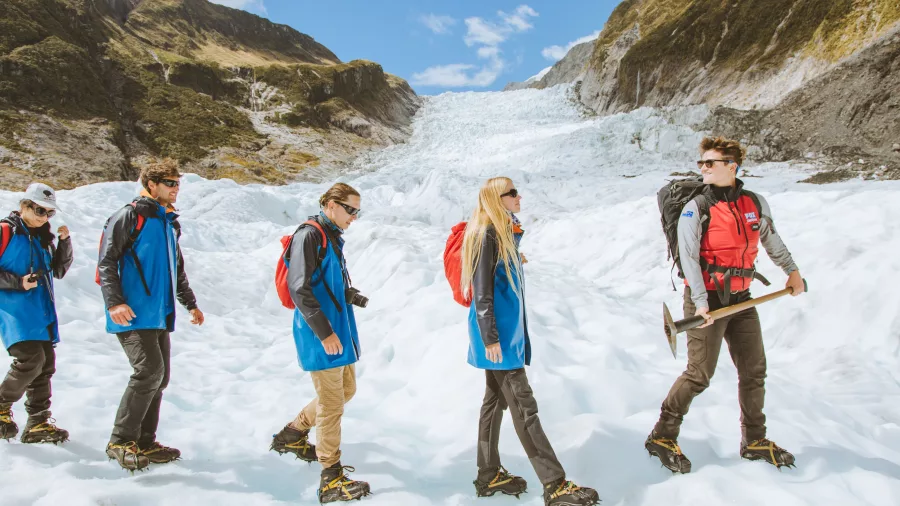 A line of hikers walks across Fox Glacier with mountain peaks rising behind.