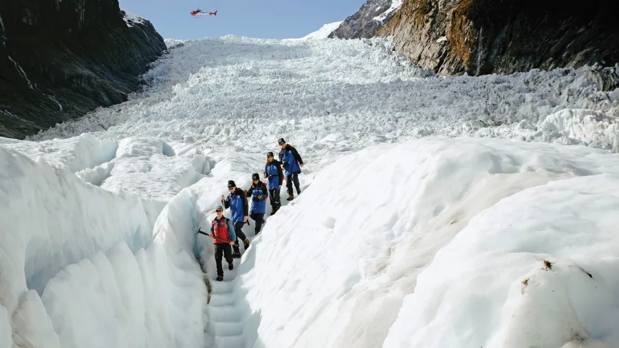 Hikers descend an ice staircase carved into the glacier, with a helicopter flying above.