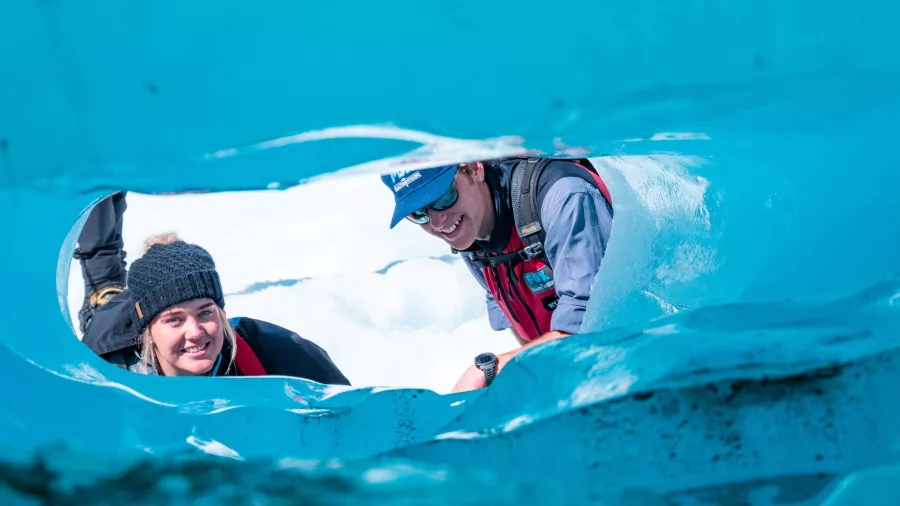 Two hikers peer through a bright blue ice tunnel on Fox Glacier.