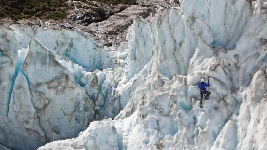 Climber scales a towering ice wall on Fox Glacier surrounded by dramatic glacial formations.