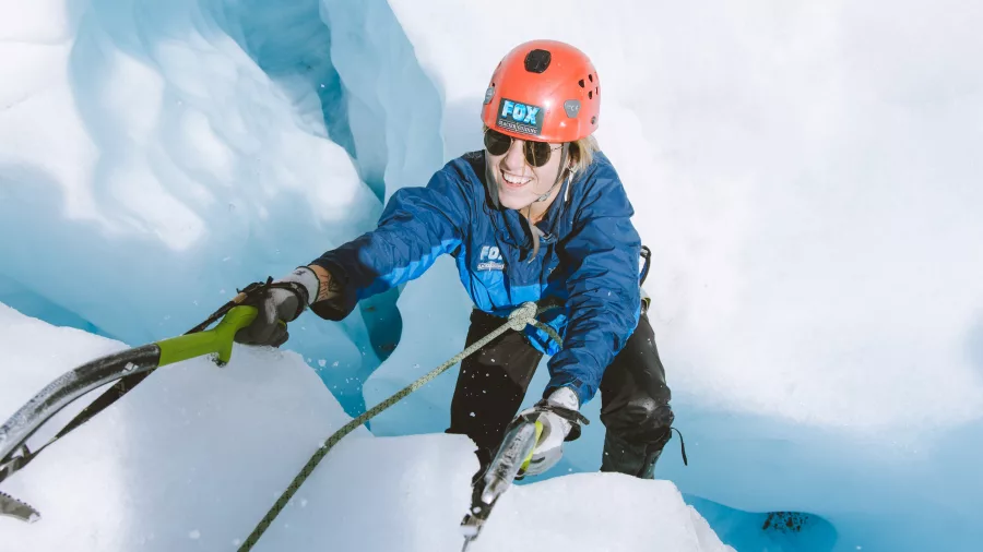 A smiling climber ascends a bright blue ice wall on Fox Glacier using ice axes.