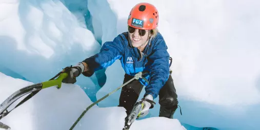 A smiling climber ascends a bright blue ice wall on Fox Glacier using ice axes.