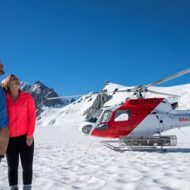 Couple standing next to a red and white helicopter on a snowy helihike tour on Fox Glacier