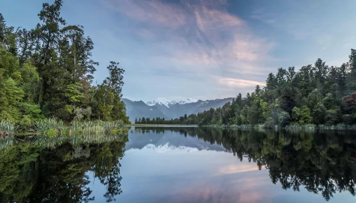 Still waters of Lake Matheson reflecting trees and distant snow-capped peaks at dusk on the West Coast of New Zealand