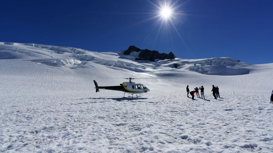 Helicopter and tourists on a snowy landing zone at Fox Glacier under a bright sun