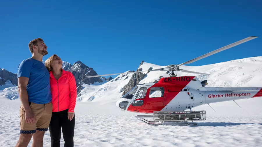 Couple standing next to a red and white helicopter on a snowy helihike tour on Fox Glacier