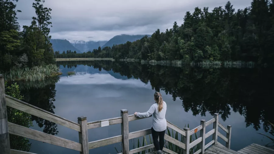 Woman standing on the wooden viewing platform overlooking the reflective waters of Lake Matheson near Fox Glacier, New Zealand