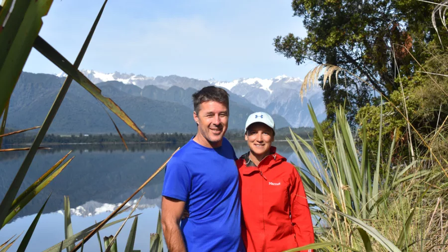 Couple standing beside Lake Mapourika with mountains and reflections behind them