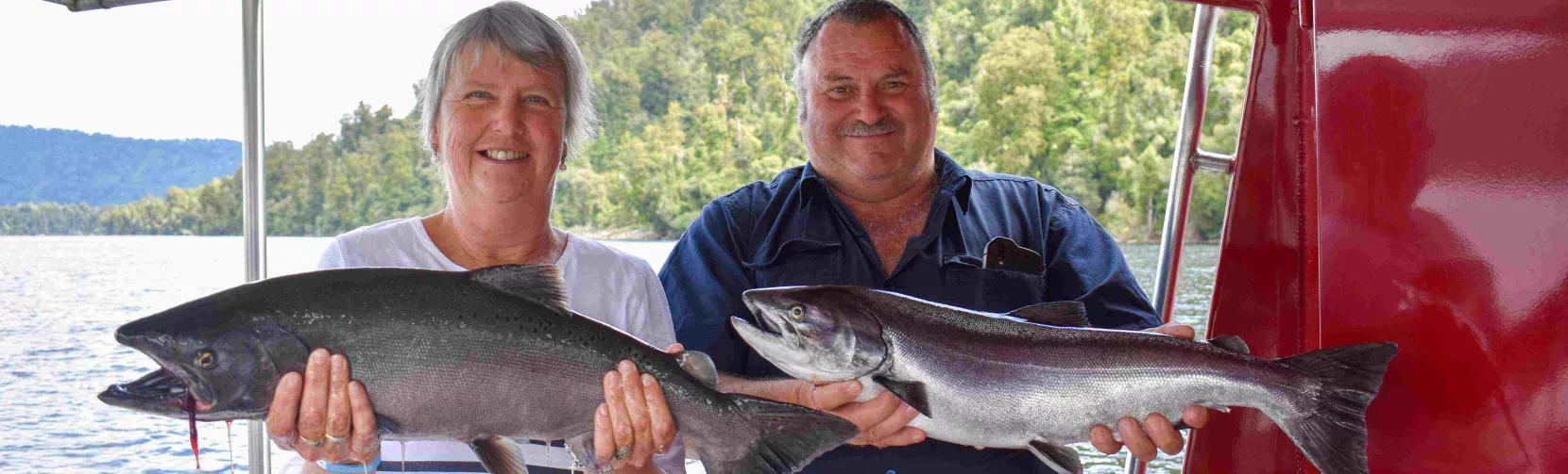 Smiling couple holding freshly caught salmon aboard a fishing boat on Lake Mapourika