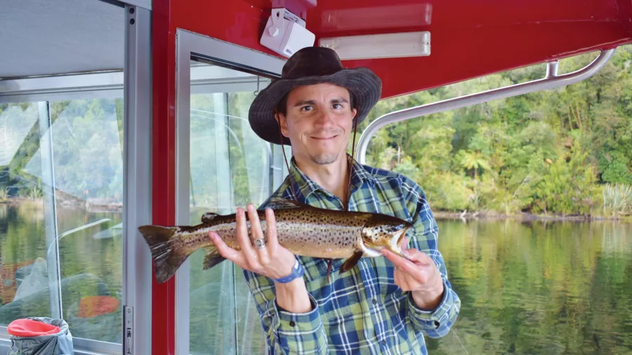 Man in a checkered shirt and hat holding a brown trout on a fishing boat in Lake Mapourika