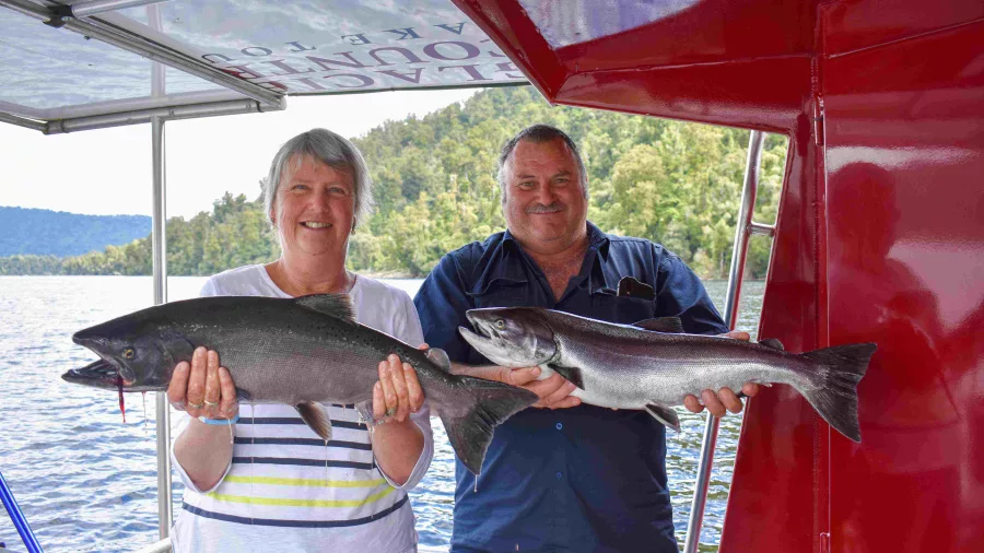 Smiling couple holding freshly caught salmon aboard a fishing boat on Lake Mapourika