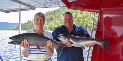 Smiling couple holding freshly caught salmon aboard a fishing boat on Lake Mapourika