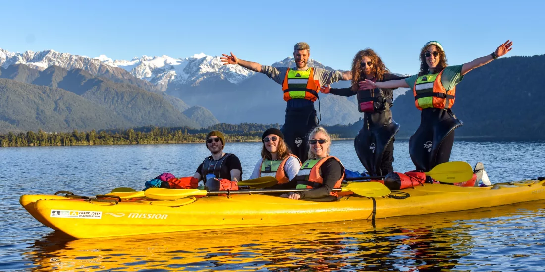 Group of people in yellow kayaks posing on a lake with snow-capped mountains in the background