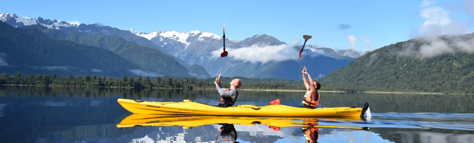 Two kayakers toss paddles in the air on a calm lake in Franz Josef with snow-capped mountains reflected in the water