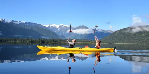 Two kayakers toss paddles in the air on a calm lake in Franz Josef with snow-capped mountains reflected in the water
