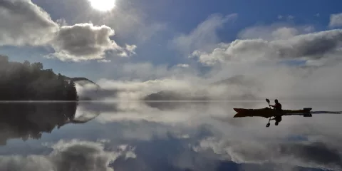 Solo kayaker silhouetted against the sun with clouds and reflections on a still lake in Franz Josef