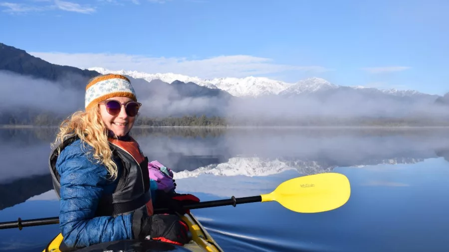 Woman kayaking on a calm lake with snow-covered Southern Alps in the background during Franz Josef Wilderness Tours’ Kayak Classic winter experience