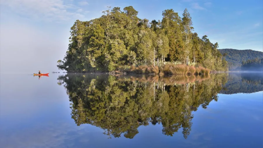 Solo kayaker paddling near a forested island with perfect reflections on the lake