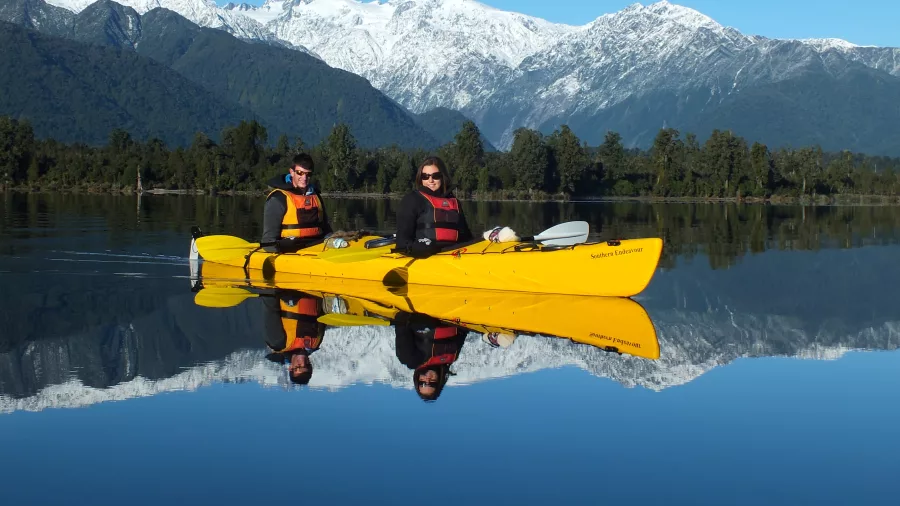 Two people in a yellow kayak on a calm lake with snowy mountains and reflections in the water