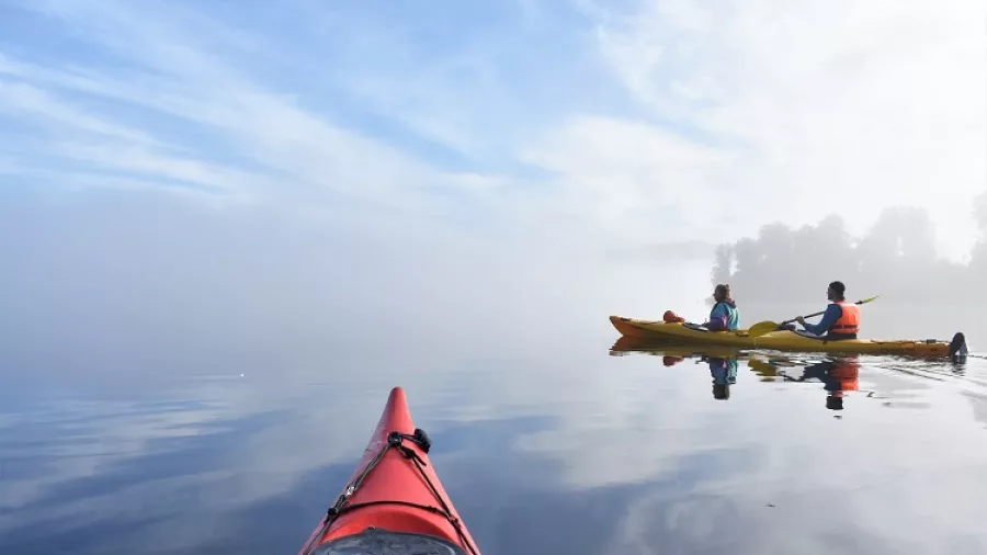 View from a red kayak following another pair across a misty reflective lake in Franz Josef
