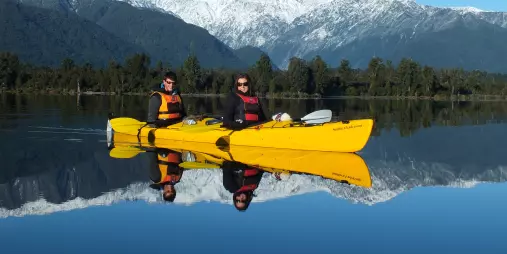 Two people in a yellow kayak on a calm lake with snowy mountains and reflections in the water