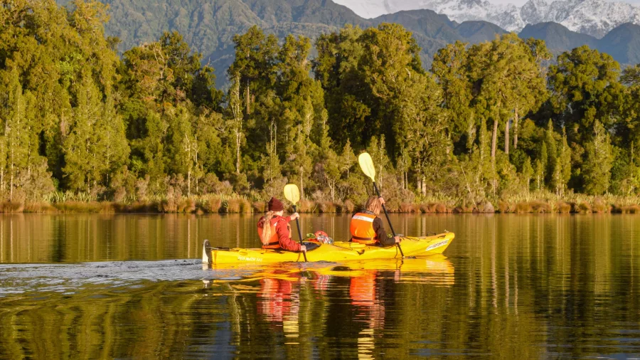 Two people kayaking on Lake Mapourika during a Franz Josef Sunset Kayaking Tour with forest reflections and snow-capped mountains in the background.