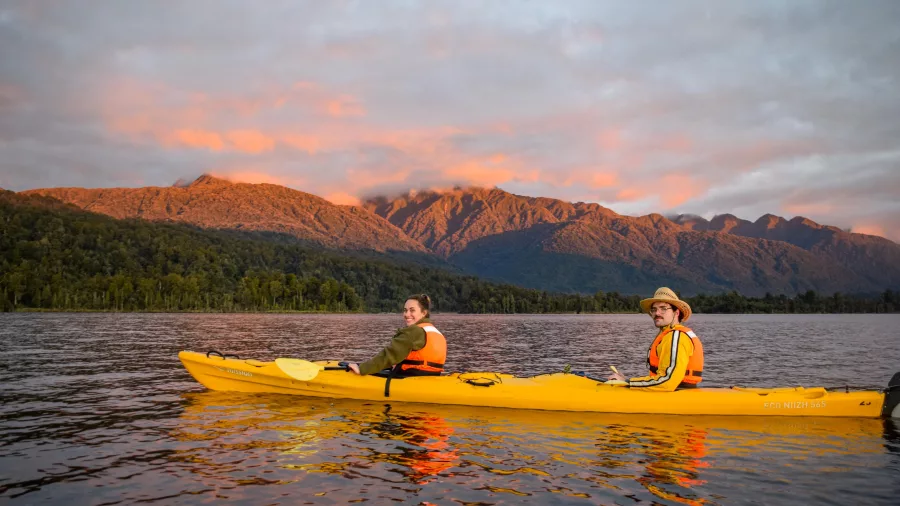 Two people kayaking on Lake Mapourika during sunset with golden light hitting the mountains near Franz Josef Glacier.