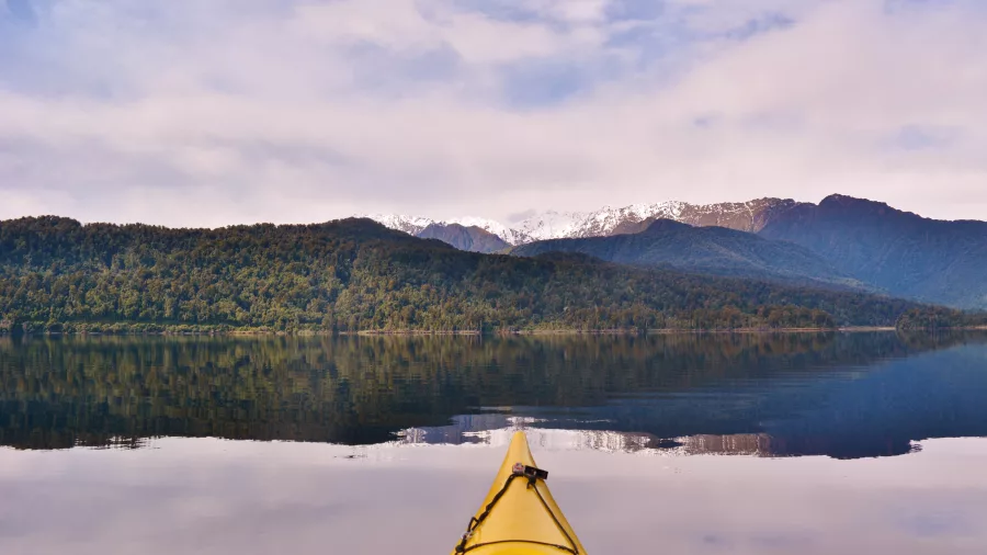 Front view from a yellow kayak on Lake Mapourika with calm reflections and snow-capped mountains near Franz Josef.