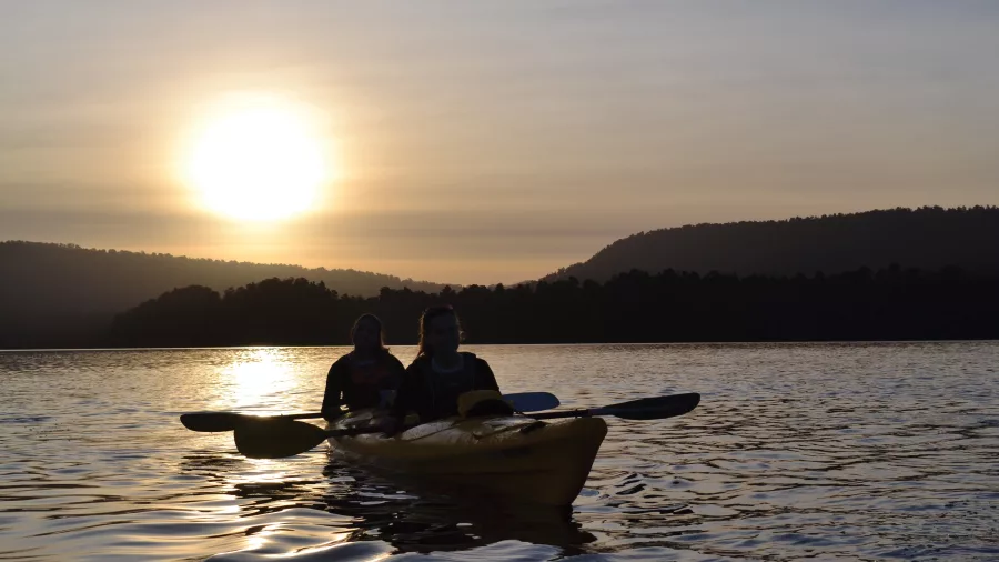 Two people kayaking on Lake Mapourika during sunset with forested hills silhouetted in the background.