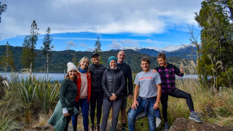 Two kayakers paddle near forested shoreline with snow-covered Southern Alps in the background