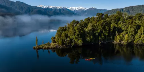 Aerial view of kayakers paddling near forested island on West Coast lake with Southern Alps in the background