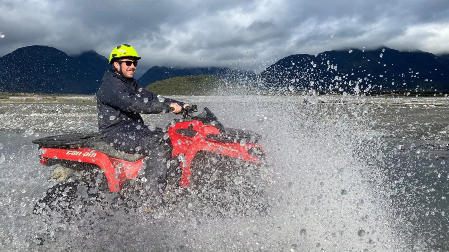 Man riding a red quad bike through a splash of water with West Coast mountains in the background