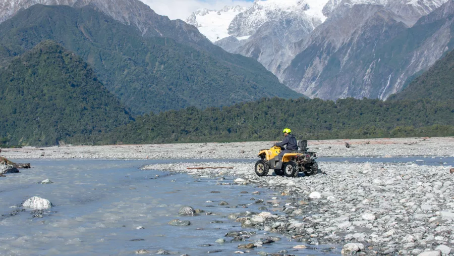 Two people riding a yellow quad bike across a rocky riverbed with glacier-covered mountains in the distance
