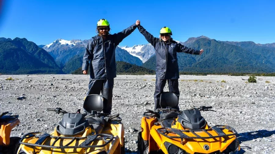Two people standing on yellow quad bikes and holding hands with snow-capped mountains in the background near Franz Josef