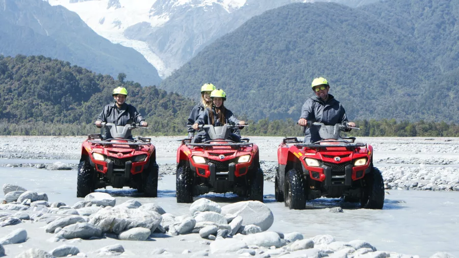 Group riding red quad bikes through shallow glacier-fed river with Franz Josef Glacier in the background