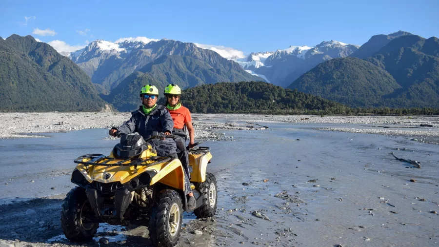 Two people on a yellow quad bike with Franz Josef Glacier and Southern Alps in the background