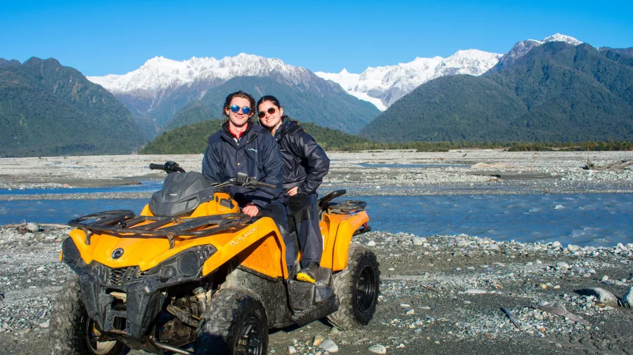 Couple posing on a yellow quad bike beside the river with Franz Josef Glacier in the background