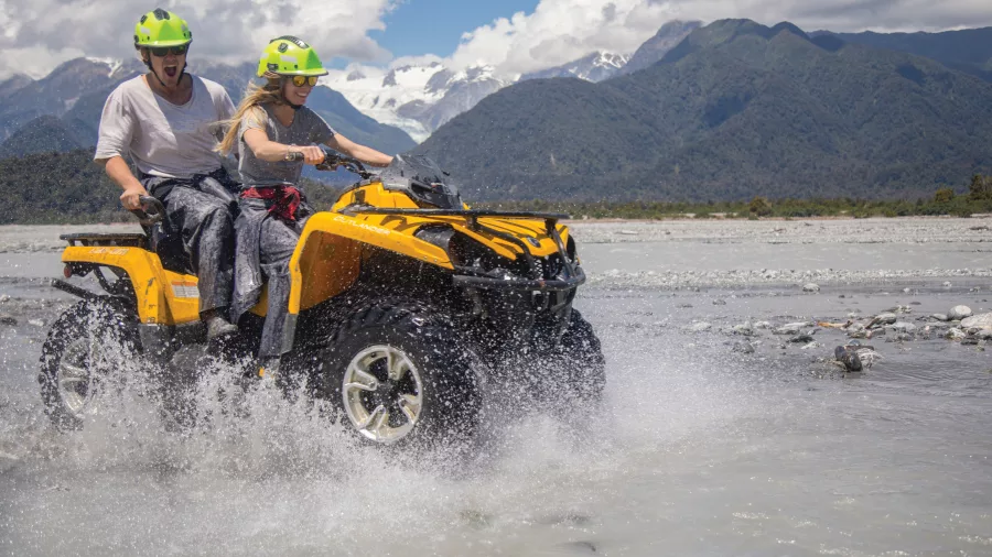 Smiling couple rides a yellow quad bike through a shallow river with Franz Josef Glacier in the background