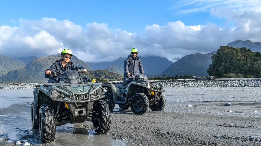 Two people riding quad bikes across a rocky riverbed on the West Coast of New Zealand