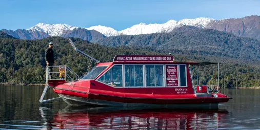 Red Franz Josef Wilderness Tours boat cruising on Lake Mapourika with snow-capped mountains in the background