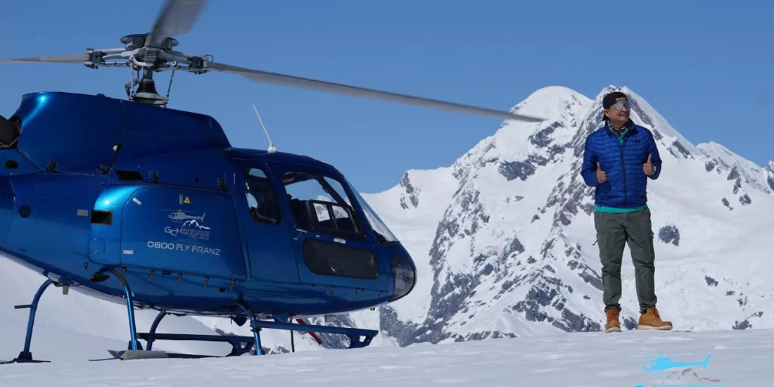 Man standing beside Glacier Country Helicopters AS350 with snow-covered Southern Alps in the background