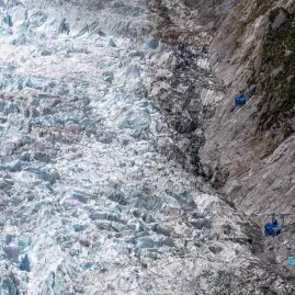 Two GCH Squirrel helicopters flying over the Franz Josef Glacier with dramatic rocky cliffs in view