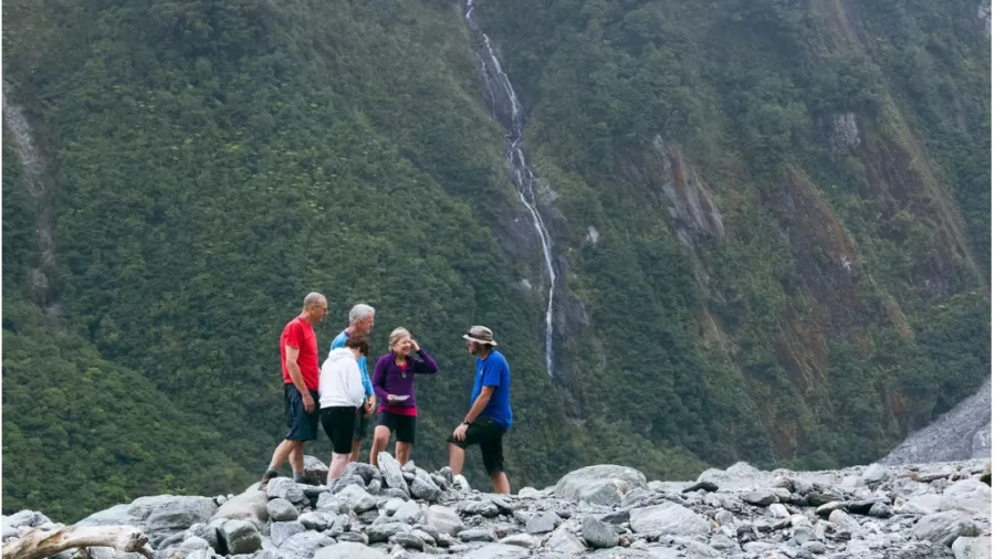 Guided group tour standing on rocks in Fox Glacier valley with lush cliffs in background