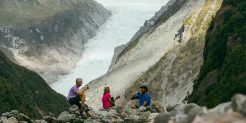 Three people enjoying a tea break with a view of Fox Glacier from the valley floor on a guided eco walk