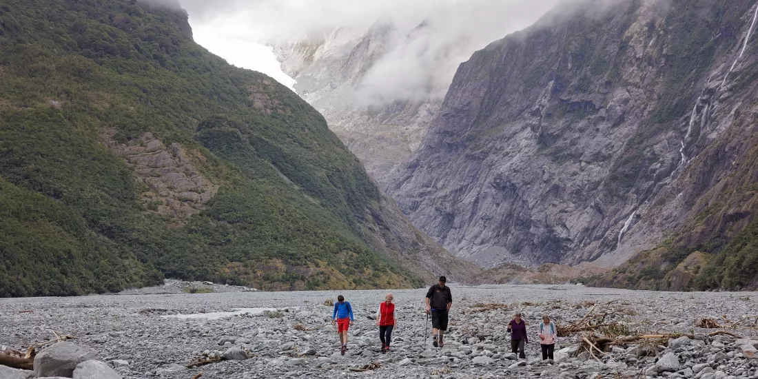 Group of walkers crossing the Franz Josef Glacier valley floor under dramatic cloud cover