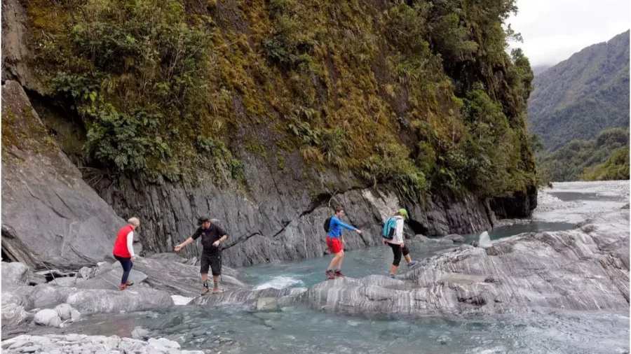 Small group crossing glacier-fed stream surrounded by steep cliffs and native forest in Franz Josef, New Zealand