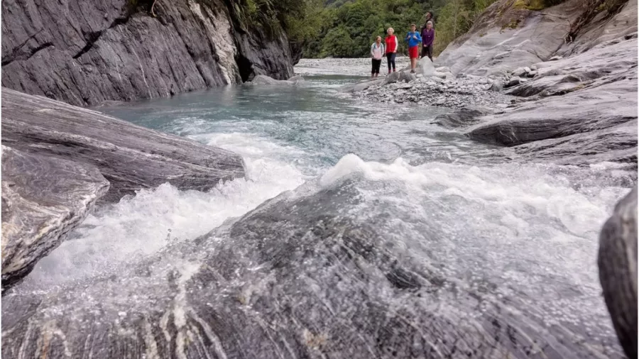 Fast-flowing glacier stream and hikers in rocky valley on New Zealand’s West Coast
