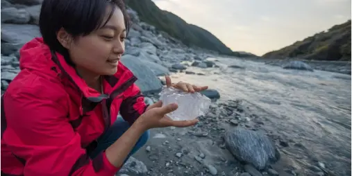Woman holding a clear chunk of glacier ice beside a rocky river in Franz Josef, New Zealand